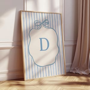 May include: A framed wall art print featuring a light blue letter "D" inside a decorative shape, set against a background of vertical blue and cream stripes. The artwork is in a wooden frame and leans against a wall.
