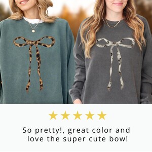 May include: Two women wearing sweatshirts with a large bow design. The bow is made of a leopard print fabric on one sweatshirt and a white and gray print on the other.