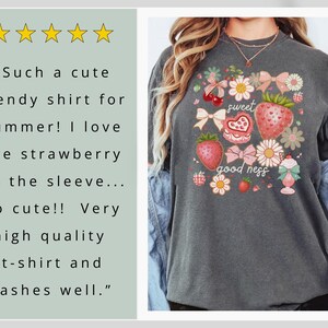 Op de afbeelding: Donkergrijs T-shirt met een aardbeienthema. Het ontwerp bevat aardbeien, strikken, bloemen en de woorden "sweet good ness". Het shirt heeft een ronde hals en korte mouwen.