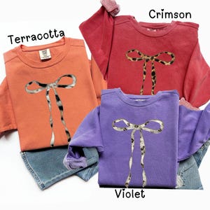 May include: Three sweatshirts with a leopard print bow design. The sweatshirts are in terracotta, crimson, and violet.