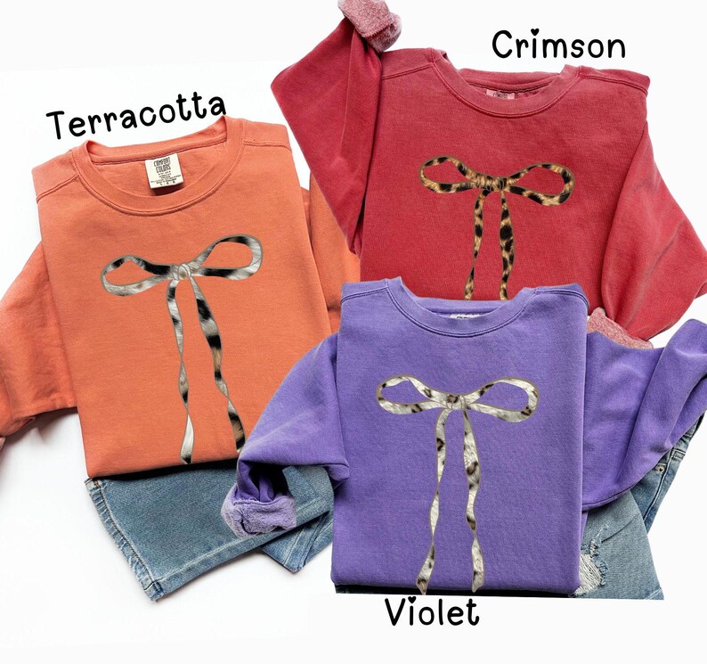 May include: Three sweatshirts with a leopard print bow design. The sweatshirts are in terracotta, crimson, and violet.