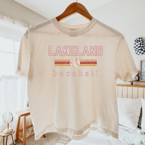 May include: Cream-colored t-shirt with the text "LAKELAND baseball" in red and yellow. A baseball graphic is centered above the word "baseball". The shirt is hanging on a wooden hanger.