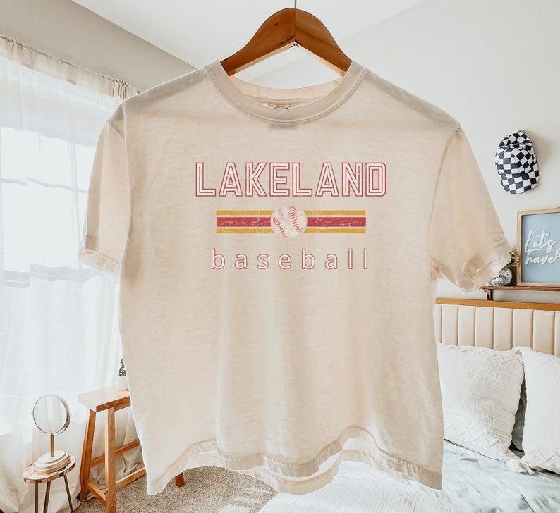 May include: Cream-colored t-shirt with the text "LAKELAND baseball" in red and yellow. A baseball graphic is centered above the word "baseball". The shirt is hanging on a wooden hanger.