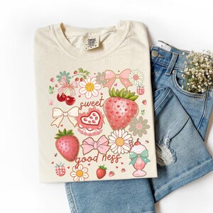 Op de afbeelding: Cr&egrave;mekleurig T-shirt met een grillig aardbeienthema met de woorden "sweet goodness". De afbeelding bevat aardbeien, strikken, bloemen en een milkshake. Het shirt wordt gecombineerd met een blauwe spijkerbroek en een klein boeket witte bloemen.