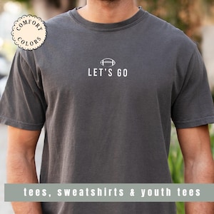May include: Dark gray t-shirt with the text "LET'S GO" and a football graphic. The shirt also features the words "Comfort Colors" in a circular design. The bottom of the shirt has the text "tees, sweatshirts & youth tees".