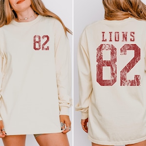 May include: Cream-colored long-sleeve shirt with the number "82" printed in distressed red on the front and back. The back of the shirt also features the word "LIONS" above the number.