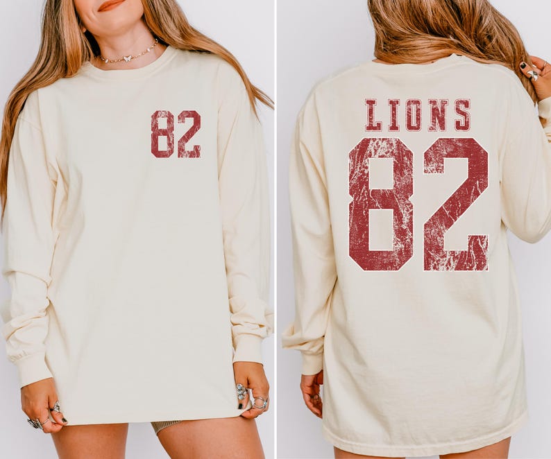 May include: Cream-colored long-sleeve shirt with the number "82" printed in distressed red on the front and back. The back of the shirt also features the word "LIONS" above the number.