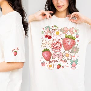 Op de afbeelding: Cr&egrave;mekleurig T-shirt met een aardbeienthema. Het ontwerp bevat aardbeien, strikken, bloemen en de woorden "sweet good ness". Een kleinere versie van het ontwerp staat op de mouw.