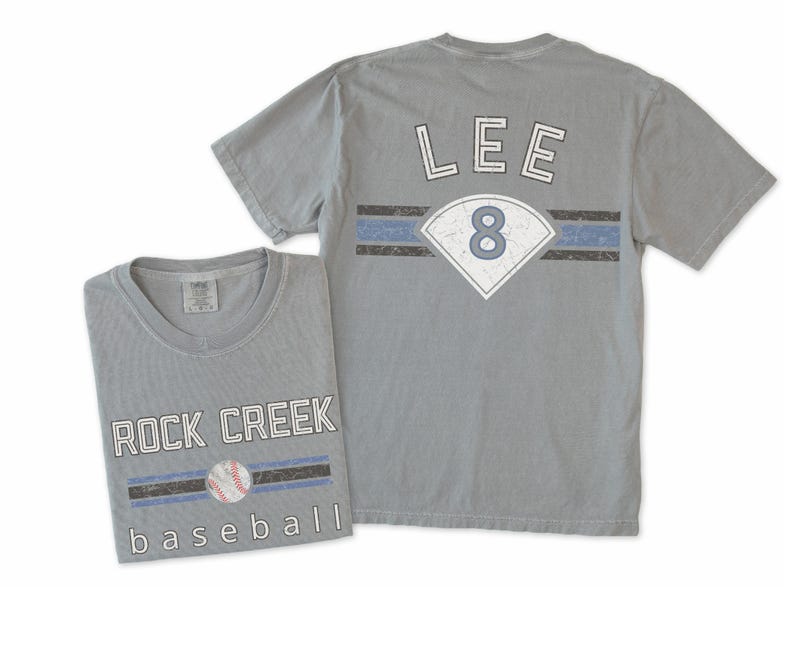 May include: Two gray baseball t-shirts. One shirt is folded, displaying "ROCK CREEK baseball" with a baseball graphic. The other shirt shows "LEE 8" on the back, with a baseball diamond graphic and blue and black stripes.