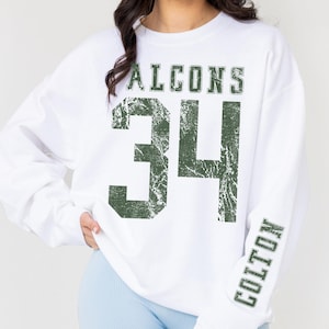 May include: White sweatshirt with the word "FALCONS" above the number "34" in distressed green lettering. The name "COLTON" is printed vertically on the right sleeve in green. The sweatshirt is a casual, sporty style.
