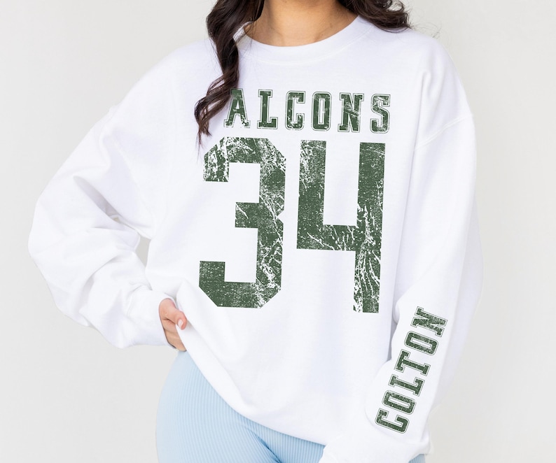 May include: White sweatshirt with the word "FALCONS" above the number "34" in distressed green lettering. The name "COLTON" is printed vertically on the right sleeve in green. The sweatshirt is a casual, sporty style.