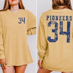 May include: Mustard yellow long-sleeve shirt with the number 34 in navy blue on the front and back. The word "PIONEERS" is printed above the number on the back. The shirt has a relaxed fit and a crew neck.