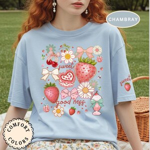 Op de afbeelding: Lichtblauw T-shirt met een aardbeienthema. Het ontwerp bevat aardbeien, bloemen, strikken en de woorden "sweet" en "good ness". Het shirt heeft korte mouwen en een ronde hals. De tekst "CHAMBRAY" staat rechtsboven.