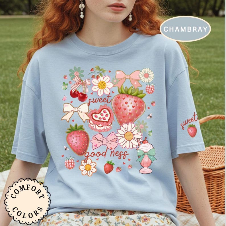 Op de afbeelding: Lichtblauw T-shirt met een aardbeienthema. Het ontwerp bevat aardbeien, bloemen, strikken en de woorden "sweet" en "good ness". Het shirt heeft korte mouwen en een ronde hals. De tekst "CHAMBRAY" staat rechtsboven.