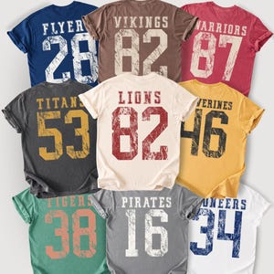 May include: A collection of vintage-style t-shirts in various colors, each featuring a team name and number. The shirts include "FLYER 28", "VIKINGS 82", "WARRIORS 87", "TITANS 53", "LIONS 82", "WOLVERINES 46", "TIGERS 38", "PIRATES 16", and "PIONEERS 34".