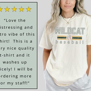 May include: Cream-colored t-shirt with the text "WILDCAT baseball" in a collegiate font, featuring a baseball graphic. The shirt has a retro design with green and gold stripes. The shirt is made of a soft material.