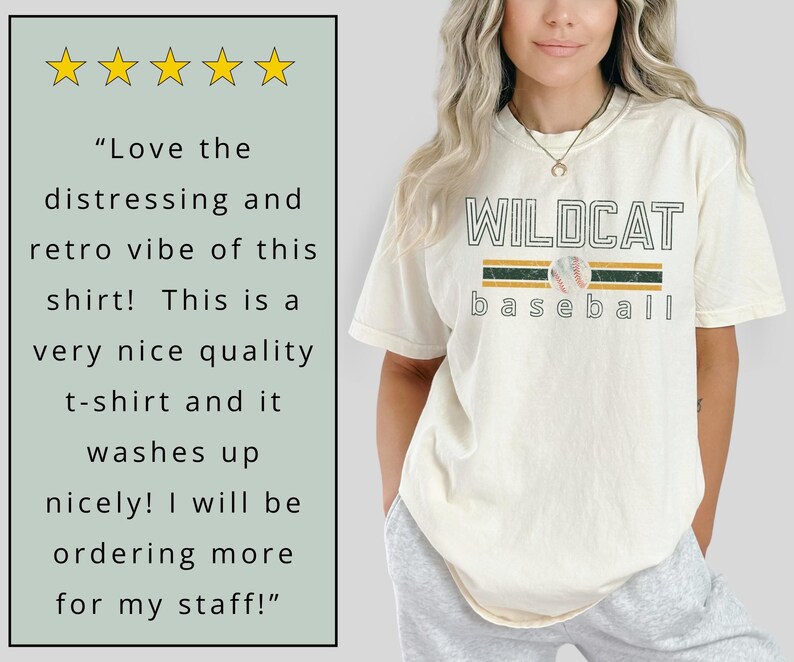 May include: Cream-colored t-shirt with the text "WILDCAT baseball" in a collegiate font, featuring a baseball graphic. The shirt has a retro design with green and gold stripes. The shirt is made of a soft material.