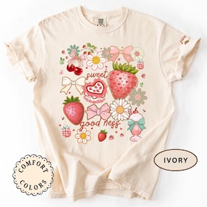 Op de afbeelding: Ivoor kleurig T-shirt met een aardbeien thema ontwerp. De afbeelding bevat aardbeien, strikken, bloemen en de woorden "sweet goodness". Het shirt is gemaakt door Comfort Colors.