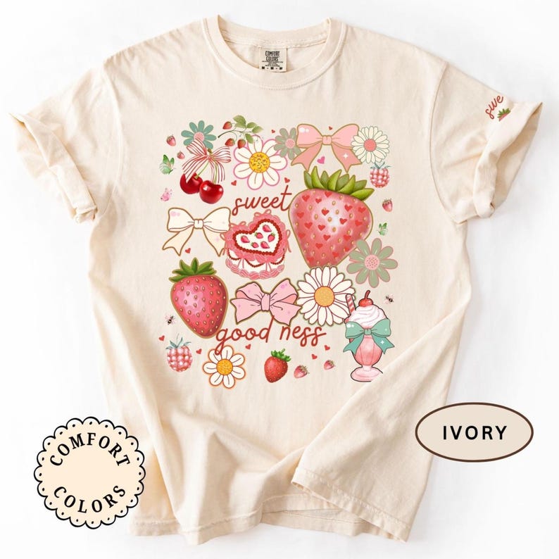 Op de afbeelding: Ivoor kleurig T-shirt met een aardbeien thema ontwerp. De afbeelding bevat aardbeien, strikken, bloemen en de woorden "sweet goodness". Het shirt is gemaakt door Comfort Colors.