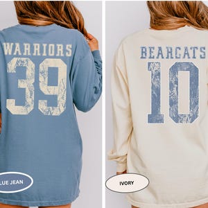May include: Two long-sleeved shirts with distressed text and numbers on the back. One is blue jean with "WARRIORS" and the number "39". The other is ivory with "BEARCATS" and the number "10". Both shirts have a vintage, worn look.