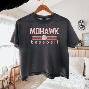 May include: Dark gray t-shirt with the text "MOHAWK baseball" in a red and white design. The word "MOHAWK" is above a baseball graphic, and the word "baseball" is below. The shirt is hanging on a wooden hanger.