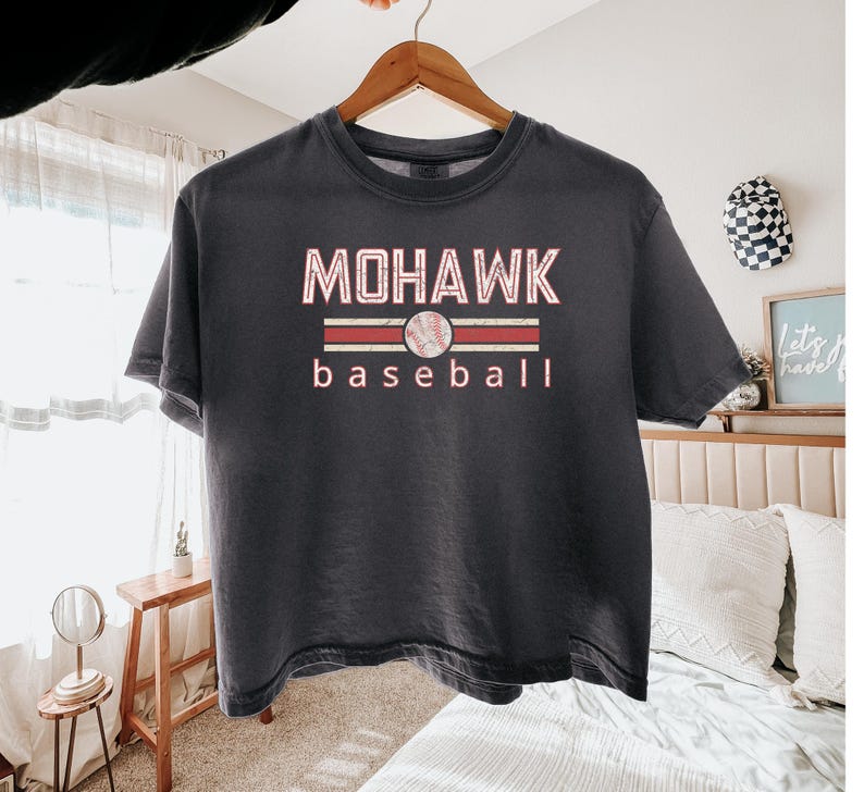 May include: Dark gray t-shirt with the text "MOHAWK baseball" in a red and white design. The word "MOHAWK" is above a baseball graphic, and the word "baseball" is below. The shirt is hanging on a wooden hanger.