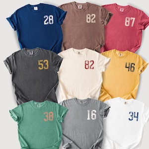 May include: Nine short-sleeved t-shirts in various colors, including navy blue, brown, and white. Each shirt features a distressed number graphic on the front. The numbers are in different colors, such as white, gold, and red.