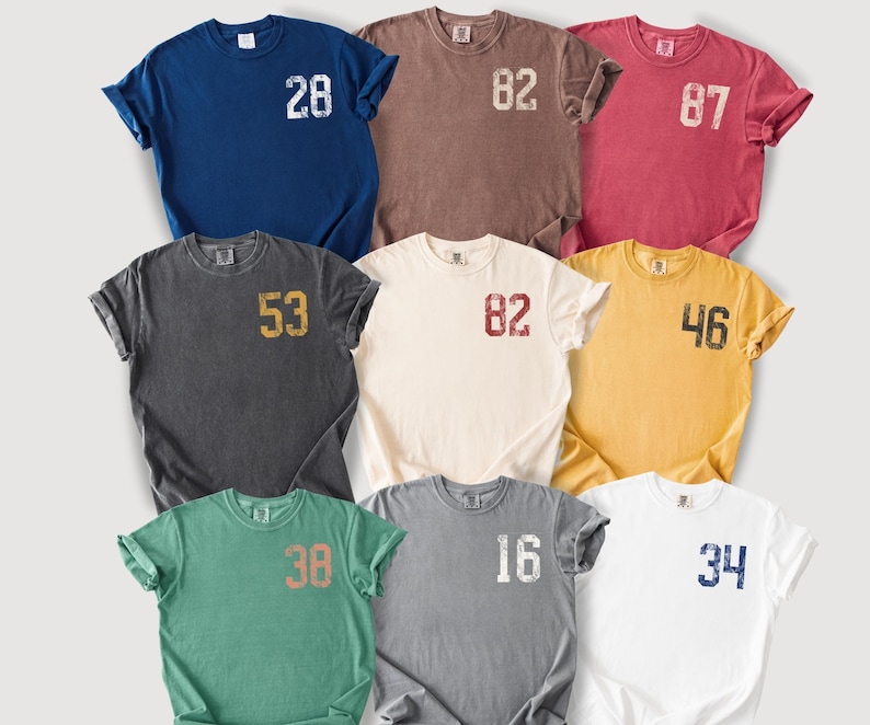 May include: Nine short-sleeved t-shirts in various colors, including navy blue, brown, and white. Each shirt features a distressed number graphic on the front. The numbers are in different colors, such as white, gold, and red.