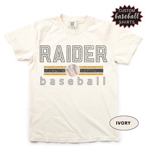May include: Ivory-colored t-shirt with the word "RAIDER" in a black, outlined font above a baseball graphic. Below the baseball is the word "baseball". The shirt has a distressed look and the text "CUSTOM BASEBALL SHIRTS" is in a starburst graphic.