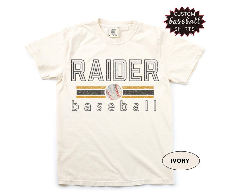 May include: Ivory-colored t-shirt with the word "RAIDER" in a black, outlined font above a baseball graphic. Below the baseball is the word "baseball". The shirt has a distressed look and the text "CUSTOM BASEBALL SHIRTS" is in a starburst graphic.