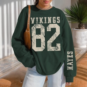 May include: A dark green sweatshirt with the word "VIKINGS" above the number "82" in a distressed, off-white font. The name "HAYES" is printed vertically on the right sleeve. The sweatshirt is a long-sleeved crew neck.
