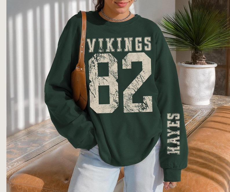 May include: A dark green sweatshirt with the word "VIKINGS" above the number "82" in a distressed, off-white font. The name "HAYES" is printed vertically on the right sleeve. The sweatshirt is a long-sleeved crew neck.