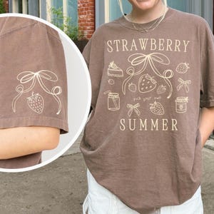 May include: A brown t-shirt with a white graphic design featuring strawberries, bows, and the words "STRAWBERRY SUMMER". The design includes illustrations of strawberry shortcake, jars, and other summer-themed elements. The left sleeve has a smaller strawberry design.