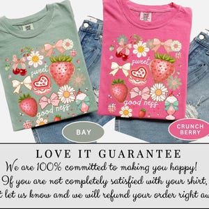 Op de afbeelding: Twee T-shirts, &eacute;&eacute;n saliegroen en &eacute;&eacute;n roze, elk met een aardbei-thema. Het ontwerp bevat aardbeien, strikken, bloemen en de woorden "sweet" en "good ness". De shirts worden gecombineerd met denim shorts.