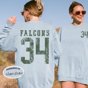 May include: Light blue sweatshirt with the word "FALCONS" and the number "34" printed in distressed green. The sweatshirt is a casual style, perfect for sports fans.