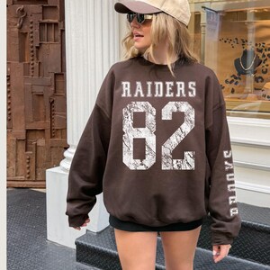 May include: A brown Raiders sweatshirt with the number 82 in white, distressed lettering. The word "RAIDERS" is above the number, and "RAIDERS" is printed vertically on the sleeve. The sweatshirt is a casual, oversized style.