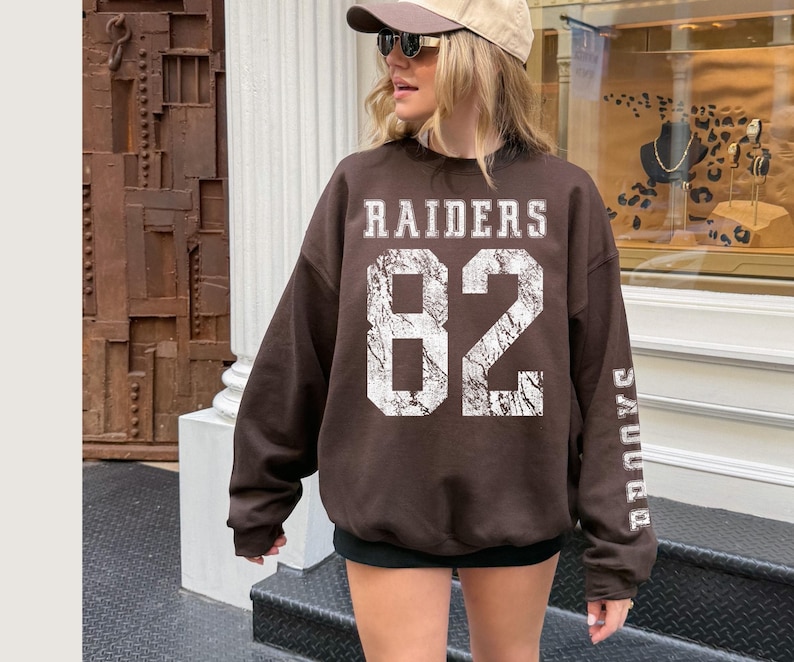 May include: A brown Raiders sweatshirt with the number 82 in white, distressed lettering. The word "RAIDERS" is above the number, and "RAIDERS" is printed vertically on the sleeve. The sweatshirt is a casual, oversized style.