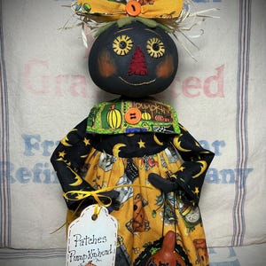 May include: A fabric doll with a round black head, a red triangular nose, and button eyes. It wears a yellow bow, a green collar with pumpkin prints, and a yellow dress featuring moon and star patterns. It holds a small pumpkin with a bat design.