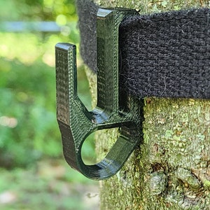 May include: A black plastic hook attached to a black strap around a tree trunk. The hook is designed to hold a rope or other item.