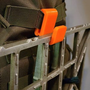 May include: A close-up of a green backpack with an orange plastic hook attached to a metal grid. The grid is made of metal bars and has a rectangular shape.