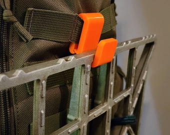 May include: A bright orange plastic J-shaped hook attached to the webbing of an olive green fabric backpack. The hook supports a metallic silver grid-like climbing stick or platform, featuring a series of notches along its top edge.