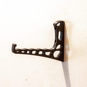 May include: A black metal wall-mounted coat rack with a series of oval cutouts along the horizontal arm. The rack has a curved design and is mounted on a white wall. The rack is designed to hold coats, hats, or other items.
