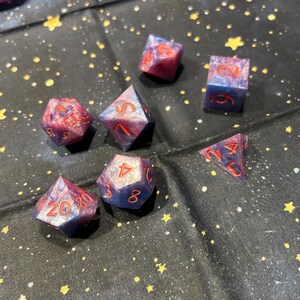 May include: A collection of seven polyhedral dice in shades of blue and red. The dice have red numbering and a marbled appearance. The dice are arranged on a black fabric with a starry pattern, suitable for tabletop gaming.