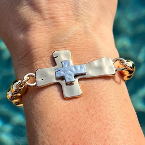 Może przedstawiać: Złota bransoletka z zawieszką w kształcie młotkowanego krzyża metalowego. Krzyż jest dwukolorowy, ze srebrnym środkiem.