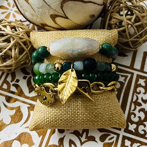 May include: A set of four bracelets featuring green agate beads, a white agate charm, a gold leaf charm, and a gold padlock charm. The bracelets are stacked on a hessian cushion.