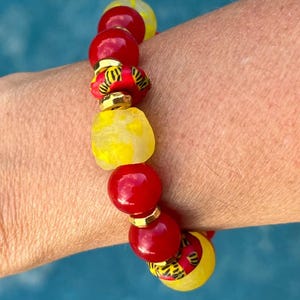 Könnte beinhalten: Ein Armband mit Perlen in Rot, Gelb und gemusterten Farben. Das Armband enthält große, runde rote Perlen, unregelmäßige gelbe Perlen und gemusterte Perlen mit goldenen Akzenten. Das Armband wird am Handgelenk getragen.