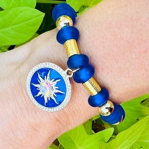Könnte beinhalten: Ein blaues und goldenes Perlenarmband mit einem Sonnenanhänger. Der Anhänger ist blau mit einer weißen Sonne und einem klaren Kristall in der Mitte. Das Armband befindet sich am Handgelenk einer Person.