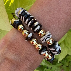 May include: Two bracelets with black and white patterned beads and gold beads. The bracelets are stacked on a person's wrist.