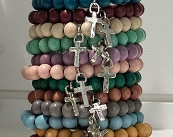 Op de afbeelding: Een stapel kleurrijke houten kralenarmbanden met zilveren kruisbedels. De armbanden zijn in tinten teal, bordeauxrood, beige, groen, paars, roze, bruin, grijs, blauw, oranje en magenta.
