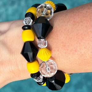 May include: A beaded bracelet featuring a combination of black, yellow, and clear beads. The bracelet includes faceted black diamond-shaped beads, round yellow beads, and clear crystal-like beads. The bracelet is worn on a wrist.